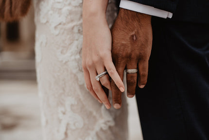 A close-up side view of a woman in white and a man in a suit holding hands and showing their wedding rings.